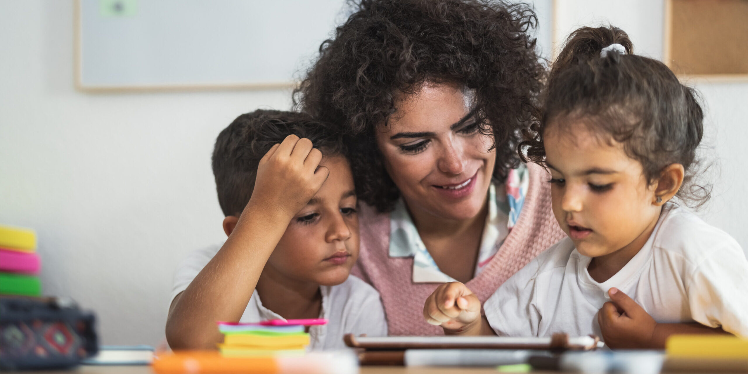 A teacher sitting with two young children, guiding them as they use a tablet in a classroom.
